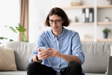 Gözlüklü neşeli bir adam kanepeye rahatça oturur ve akıllı telefonuna odaklanır. Doğal ışık modern oturma odasını dolduruyor ve boş zamanlarının tadını çıkarırken sıcak bir atmosfer yaratıyor..