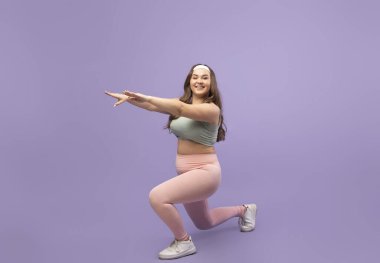 Gülümseyen, kilolu, genç bir kadın mor bir arka planda spor kıyafetleriyle hamleler yapıyor. Bu egzersiz vücut bakımını, kilo vermeyi ve kardiyo çalışmasıyla sağlıklı bir yaşam tarzını teşvik ediyor..