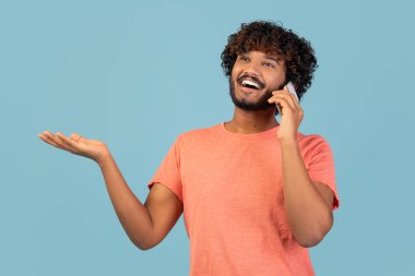 Bin yıllık neşeli, kıvırcık saçlı, telefonda konuşan Hindu bir adamın yakın çekimi. Mavi stüdyonun arka planında gülümserken heyecanla el kol hareketi yapar..