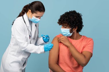 Maske takan genç bir Hintli adam mavi bir stüdyoda çalışan bir kadın sağlık görevlisinden koronavirüs aşısı alıyor. Sahne aşının önemini vurguluyor..