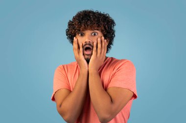 Kıvırcık saçlı, sakallı doğulu bir adam iki eliyle yüzüne dokunuyor. Ağzı çığlıkta açık, yoğun korkuyu yansıtıyor. Mavi arka plan dramatik etkiyi artırır.