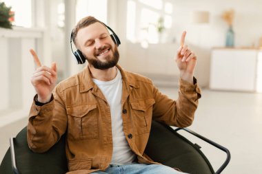 Sakallı bir adam şık bir sandalyede otururken kulaklık takar ve gülümser. Bitkilerle dolu, aydınlık bir alanda müzik dinlerken el kol hareketi yapar..