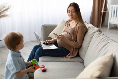 Hamile bir kadın dizüstü bilgisayar kullanarak kanepede otururken küçük bir çocuk yakınlardaki oyuncak arabalarla oynuyor. Güneş ışığı odayı doldurur, sıcak ve davetkar bir atmosfer yaratır..