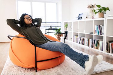 Genç bir kadın parlak turuncu bir sandalyede yaslanırken gülümsüyor. Bitkiler ve kitaplarla dolu rahat bir yaşam alanında rahat ve mutlu görünüyor. Pencereden güneş ışığı akıyor..