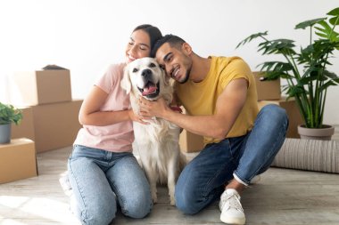 Genç bir çift, Golden Retriever 'larına sarılırken gülümseyerek yerde oturuyor. Etrafları karton kutularla çevrili. Parlak bir karşılama odasında..