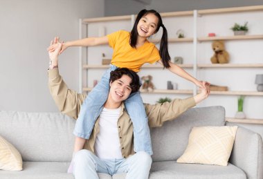 Bir baba ve kızı, rahat oturma odalarında eğlenceli etkileşimlerden hoşlanırlar. Kız, babasının omuzlarında gülerken neşeyle ışıldıyor..