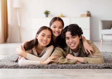 Üç genç aile ferdi, açık renkli dekorları ve basit mobilyaları olan parlak bir oturma odasında yumuşak bir halının üzerinde uzanarak birlikte kaliteli vakit geçirmenin keyfini çıkarıyorlar..