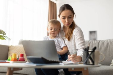 Bir anne çocuğunu kucağında tutarken dizüstü bilgisayardaki işine odaklanır. Oturma odasındalar. Masada oyuncaklar var. Günlük aile yaşantılarını sergiliyorlar..
