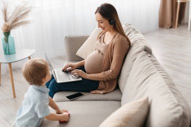 Genç bir çocuk yakınlarda oynarken hamile bir kadın dizüstü bilgisayarına odaklanmış bir şekilde kanepeye oturur. Sıcak oturma odasında yumuşak ışıklandırma ve dekoratif bitkiler var..