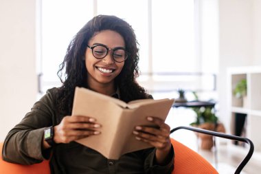 Genç bir kadın turuncu bir sandalyede rahatça oturur ve büyük bir gülümsemeyle kitabının tadını çıkarır. Doğal ışık aydınlık odayı doldurur, okumak için sıcak bir atmosfer yaratır..