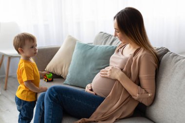 Hamile bir anne, bir oyuncak kamyonla oynayan genç oğluyla meşgul olurken bir kanepede oturur. Yumuşak gün ışığı odayı doldurur, sıcak bir atmosfer yaratır..