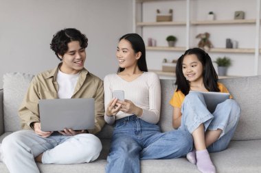 Üç genç arkadaş rahat bir oturma odasında kanepede oturup dizüstü bilgisayar, akıllı telefon ve tablet kullanırken gülümsemelerini paylaşıyorlar. Atmosfer neşeli ve rahat, mükemmel bir zaman geçirme anı..