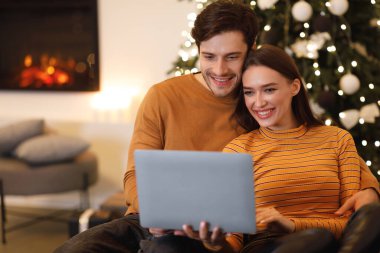 Genç bir çift kanepede yan yana oturur ve dizüstü bilgisayara bakarken gülümserler. Sıcak ortam, süslü bir Noel ağacı ve şömine ile güçlendirilir..