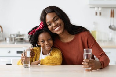 Neşeli bir anne ve kızı mutfak masasında oturmuş, ellerinde su bardaklarıyla. Gülümsemeleri ve sıcaklığı paylaşıyorlar. Parlak, neşeli bir ortamda..