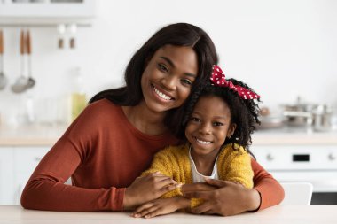 Bir anne ve kızı mutfaktaki masada birlikte oturuyorlar. Anne, her ikisi de parlak bir şekilde gülümserken kolunu kızına dolar. Mutfak sıcak ve davetkar..