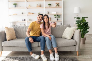 Genç bir çift, modern bir yaşam alanındaki yumuşak bir kanepede rahatça oturur, gülümser ve uzaktan kumanda tutarlar. Sıcak atmosfer bitkiler ve dekoratif raflar tarafından geliştirilir..