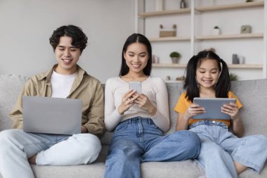 Üç genç kişi, çeşitli cihazlar kullanarak bir koltukta rahatça otururlar. Atmosfer rahat ve samimi. Parlak oturma odasındaki modern aile dijital etkileşimini yansıtıyor..