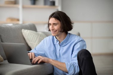 Genç bir adam şık bir oturma odasında, dizüstü bilgisayarında mutlu bir şekilde yazıyor. Doğal ışık boşluğu doldurur, görevine odaklanırken sıcak ve davetkar bir atmosfer yaratır..