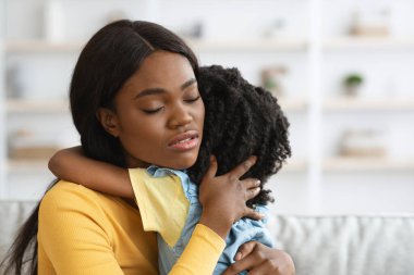 Bir anne parlak bir oturma odasında çocuğunu sıcak bir şekilde kucaklar. Duygular ve rahatlıkla dolu, sevgi dolu bir anı paylaştıklarından yakınlıkları çok açık..