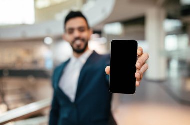 Çağdaş bir ofis ortamında takım elbiseli bir adam elinde boş bir ekranla akıllı telefonu tutarken gülümsüyor. Parlak ortam profesyonel bir atmosfer sağlıyor..