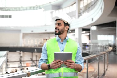 Parlak güvenlik yeleği ve sert şapkalı bir inşaat işçisi modern bir binada duruyor, tabletteki bilgileri incelerken gülümsüyor, şantiyeyi denetliyor..