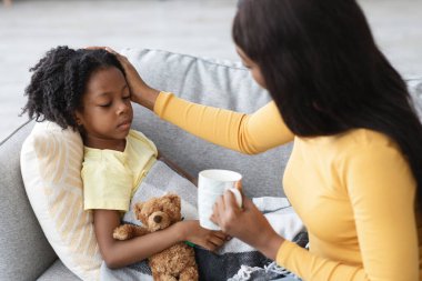 Kıvırcık saçlı bir çocuk, elinde oyuncak ayıyla kanepede dinleniyor. Bir anne nazikçe başını okşar, bir fincan çay tutarak, sıcak bir bakım atmosferi oluşturur..