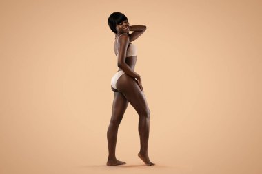 Renkli bir stüdyo ortamında, kendine güvenen bir Afro-Amerikan modeli bej iç çamaşırlarıyla şeklini sergiliyor. Çekici duruşu fit vücudunu ve uzun bacaklarını oyuncu bir şekilde vurguluyor..