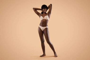 Mutlu bir Afrikalı genç Amerikalı kadın ellerini başının arkasına koyup kameraya gülümsüyor. Bej bir iç çamaşırıyla parlak bir arka plana karşı duruyor, kendine güvenini ve neşesini gösteriyor..