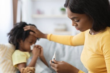 Bir anne çocuğunun sıcaklığını termometreyle ölçmeye odaklanır. Bu sırada çocuk kanepede dinlenir, hasta görünür ve elinde bir mendil tutar..