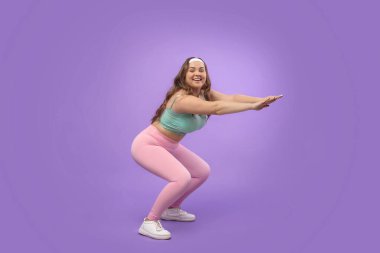 Mutlu, genç Avrupalı ve spor giyimli iri yarı bir kadın canlı, mor bir arkaplana karşı çömelme gösterisi yapıyor. Bacağını güçlendirmek ve kilo vermek için spor rutinine odaklanıyor..