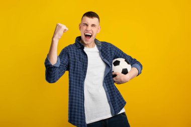 Kısa saçlı genç bir adam bir futbol topu tutarken bağırarak ve yumruğunu kaldırarak sevinç ifade eder. Canlı sarı bir arkaplanın önünde duruyor, oyuna olan hevesini gösteriyor..