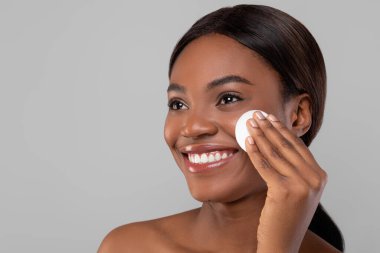 Yumuşak ciltli bir kadın pamuk ped kullanarak cilt bakım ürünü uygularken gülümsüyor. Saçları şekilli ve arka planı basit ve açık..