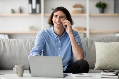 Uzun saçlı genç bir adam, parlak, modern bir oturma odasında dizüstü bilgisayarla koltukta otururken telefonla konuşurken gülümsüyor. Yakınlardaki masada bir kahve fincanı var..