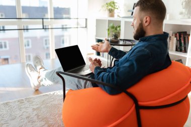 Turuncu bir sandalyede rahatça otururken bir adam sohbete dalıyor. Şehir manzarası olan geniş pencereli modern bir dairede dizüstü bilgisayar kullanıyor..