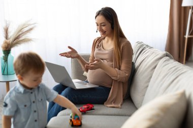 Hamile bir kadın bir kanepede oturup dizüstü bilgisayarda çalışır, gülümser ve küçük çocuğuyla arabalarla uğraşır. Güneş ışığı pencereden süzülerek sıcak bir atmosfer yaratır..