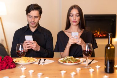 İki kişi romantik bir yemek masasında oturuyor, her biri yemek yerine akıllı telefonlarına odaklanıyor. Tabaklarında makarna ve yanında şarap bardakları var..
