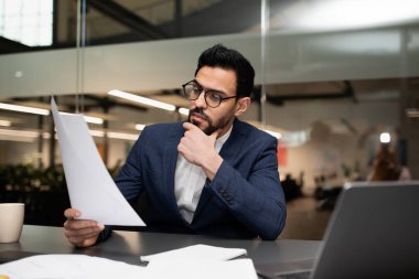 Gözlüklü ve takım elbiseli bir adam masada otururken belgeleri inceliyor. Bir dizüstü bilgisayar açık ve parlak bir ofis ortamında notlar alıyor..