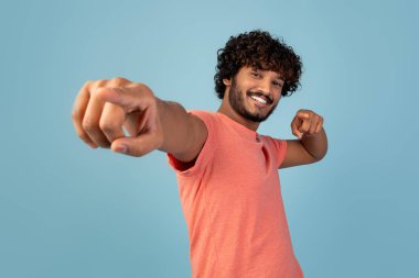 Kıvırcık saçlı ve sakallı neşeli bir Hintli genç adam mavi arka planda tek başına poz veriyor. Pembe bir tişört giyer ve kameraya dostça bir gülümsemeyle bakar..