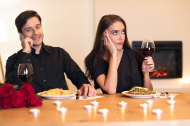 Bir çift mum ve güllerle süslenmiş bir masada oturuyor. Akşam yemeği yiyorlar, ama bir partnerin elinde bir kadeh şarap tutarken dikkati dağılmış görünüyor, potansiyel ilişki gerginliğini vurguluyor..