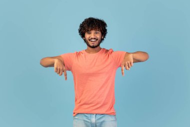 Kıvırcık saçlı neşeli bir Hintli adam gülümsüyor ve iki eliyle işaret ediyor. Mavi bir arkaplanda rahat bir şekilde poz veriyor, canlı ve çekici bir atmosfer yaratıyor..