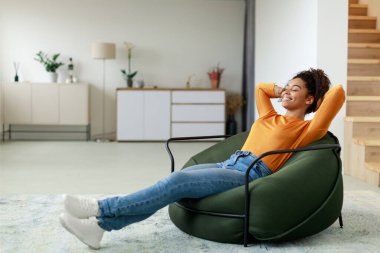 Genç bir kadın yeşil bir sandalyede eğleniyor, şık bir oturma odasında dinlenirken gülümsüyor. Parlak alan, ahşap merdivenler ve yumuşak bir dekor içeriyor, sakin bir atmosfer yaratıyor..