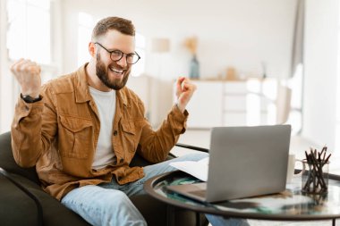 Gözlüklü neşeli bir adam rahat bir sandalyeye oturur ve kutlama için yumruklarını kaldırır. Dizüstü bilgisayarıyla meşgul, şık ev ofisinde bir anlık başarının tadını çıkarıyor..