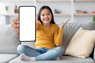 Uzun saçlı genç bir kız kanepede bacak bacak üstüne atmış, elinde boş bir ekranla akıllı bir telefon tutuyor. Parlak bir şekilde gülümsüyor ve diğer eliyle hareketler yapıyor, neşeli bir atmosfer yaratıyor..