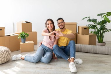 Mutlu genç çift yeni taşınmış bir oturma odasında yerde oturuyor. Elleriyle karton kutular ve bitkilerle çevrili bir kalp yapıyorlar..