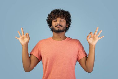 Kıvırcık saçlı genç bir Hintli pembe tişört giyer ve elleri ve gözleri kapalı meditasyon yapar. Yumuşak, mavi bir arka planda sakince oturuyor, huzuru somutlaştırıyor..