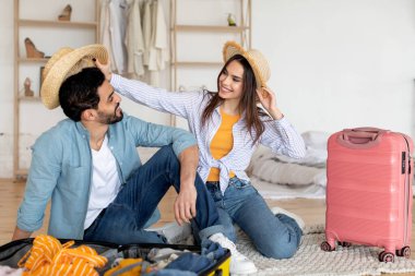 Bir çift yolculuk için toplanırken neşeli anları sever. Eğlenceli şapkalar takıp, açık bir bavul dolusu elbisenin yanında gülücüklerini paylaşıyorlar..