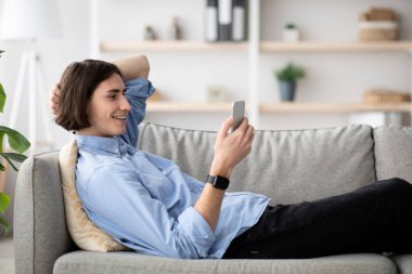 Genç bir adam şık bir oturma odasında akıllı telefonuyla meşgul olurken bir kanepede rahatça yaslanıyor. Güneş ışığı pencereden süzülerek rahat bir atmosfer yaratır..