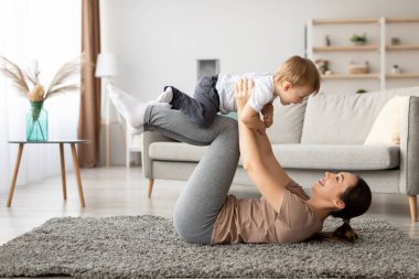 Bir anne, parlak oturma odalarında yumuşak bir halıda oynarken küçük oğlunu neşeyle kaldırır. Güneş ışığı uzayı aydınlatır ve sıcak bir atmosfer yaratır..