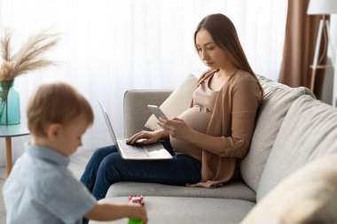 Hamile bir kadın kanepede dizüstü bilgisayarını ve akıllı telefonunu kullanarak oturur. Bu sırada da renkli oyuncaklarıyla oturma odasında dikkatle seyreder..