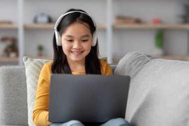 Uzun saçlı genç bir kız çevrimiçi öğrenme seansına katılırken kulaklık takıyor ve gülümsüyor. Oturma odasındaki kanepede rahatça oturuyor..